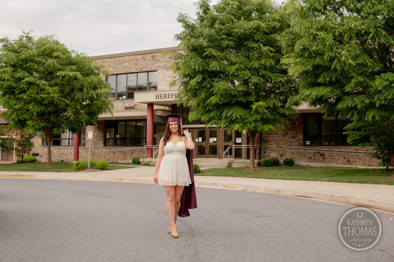 Graduate walking in white dress and burgundy gown outside brick school building.