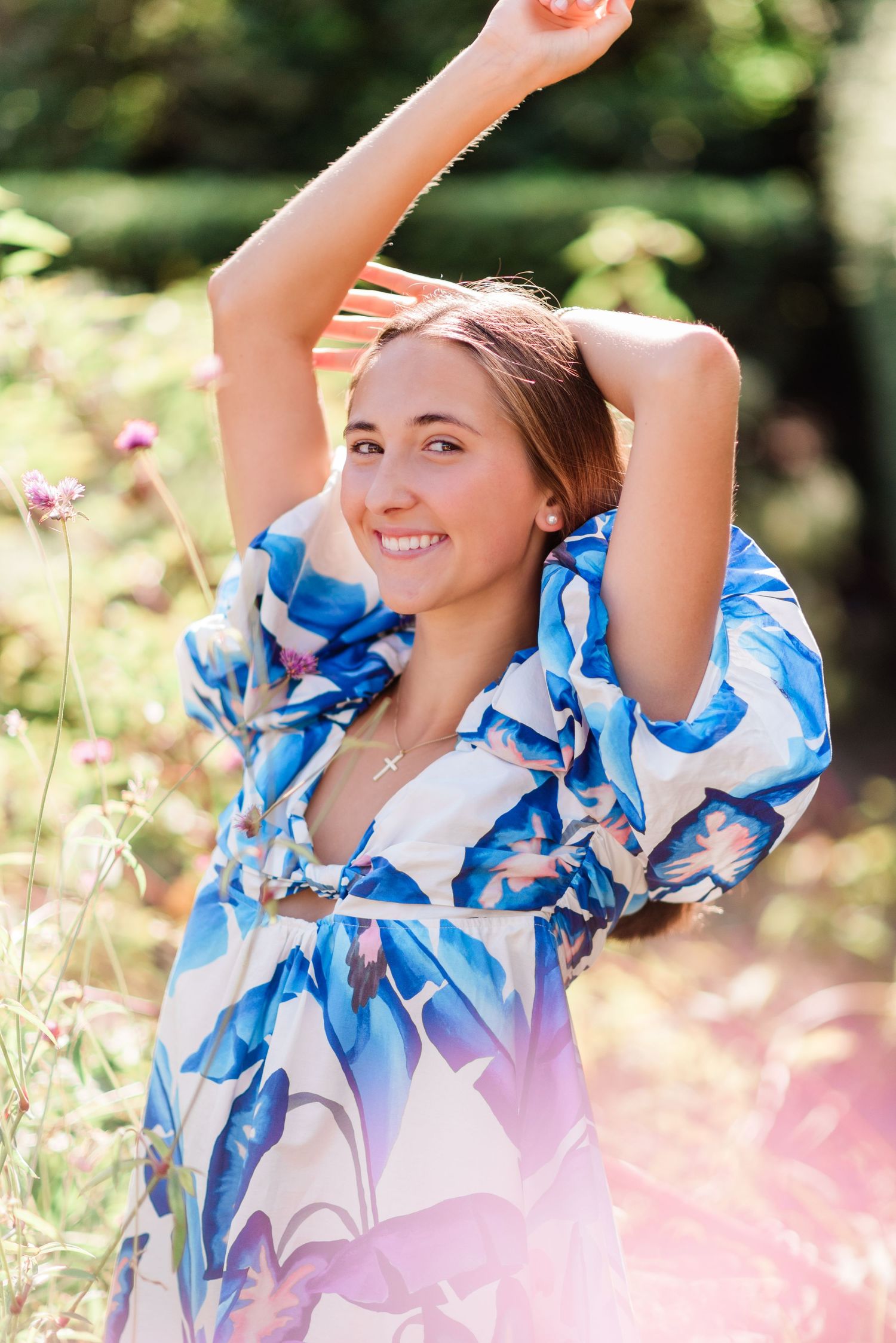 A person in a flowing blue and white floral dress poses joyfully outdoors in nature with arms raised.