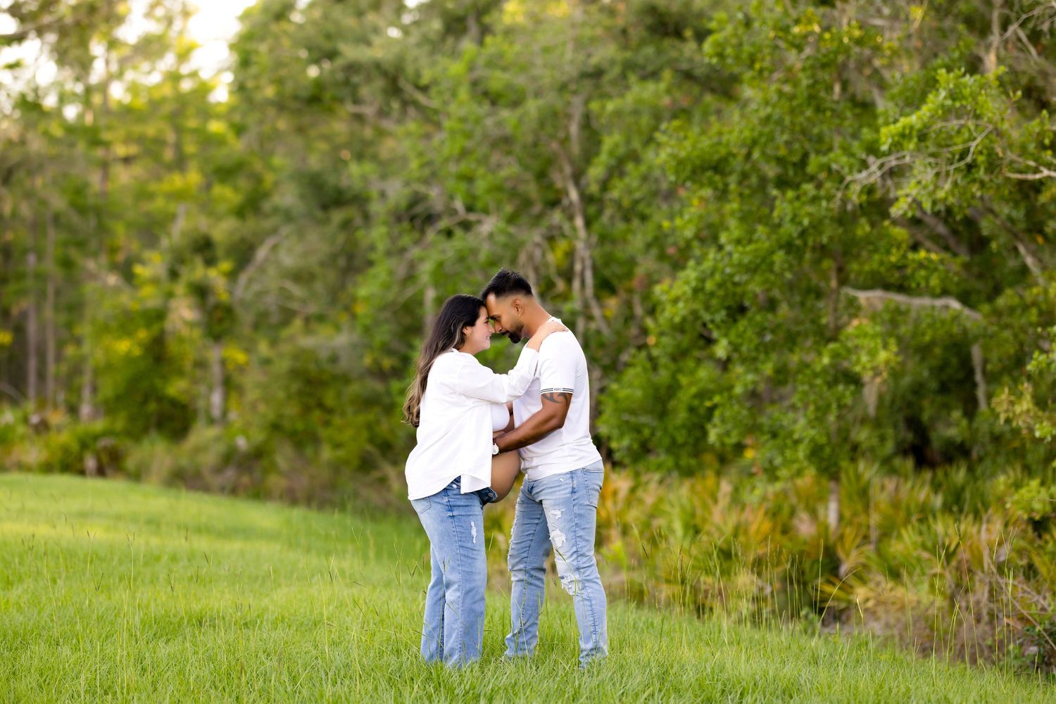 Couple standing close together in grassy field surrounded by trees.