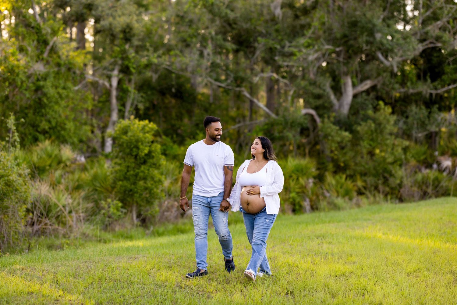 Couple strolling through grassy field with forest in background.
