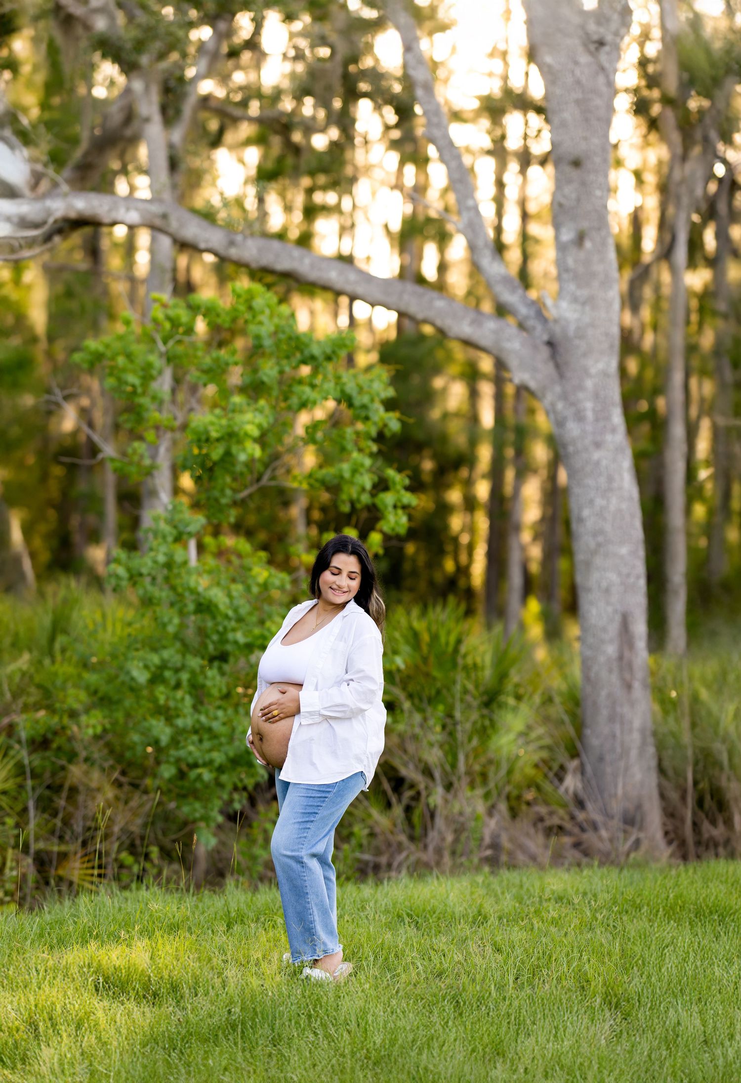Pregnant woman posing in white shirt and jeans in outdoor setting with trees.