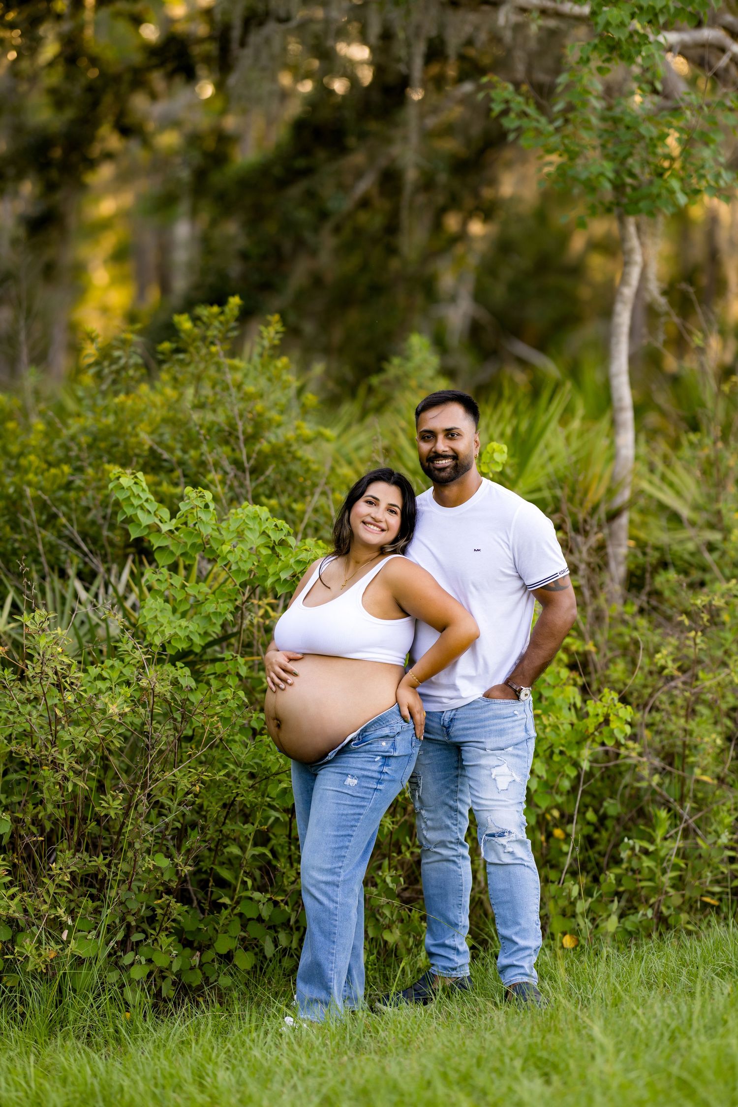 A maternity photoshoot in a lush green outdoor setting captures tender moments between a couple in matching white and denim.