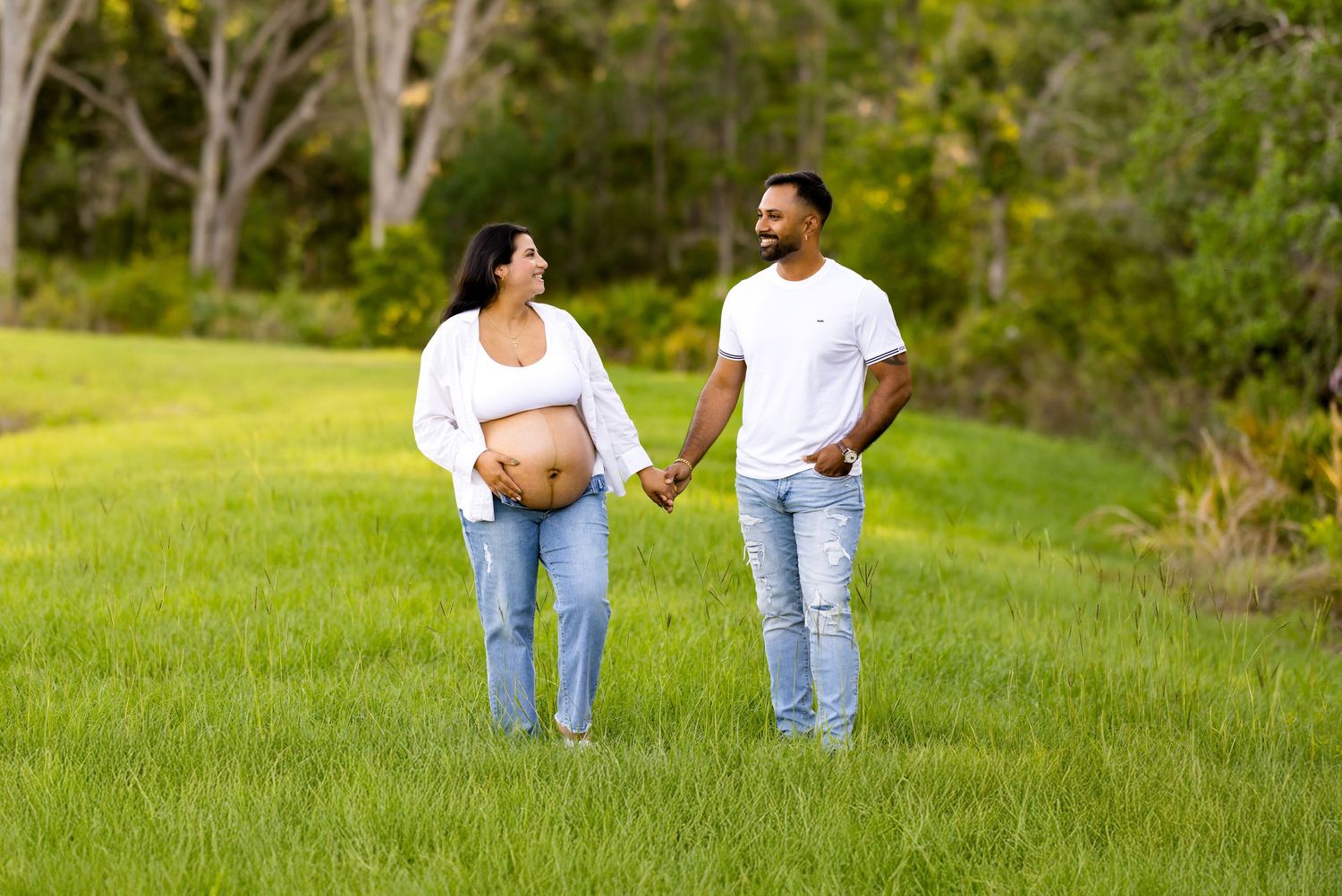 Couple walking hand-in-hand through grassy field with trees behind.