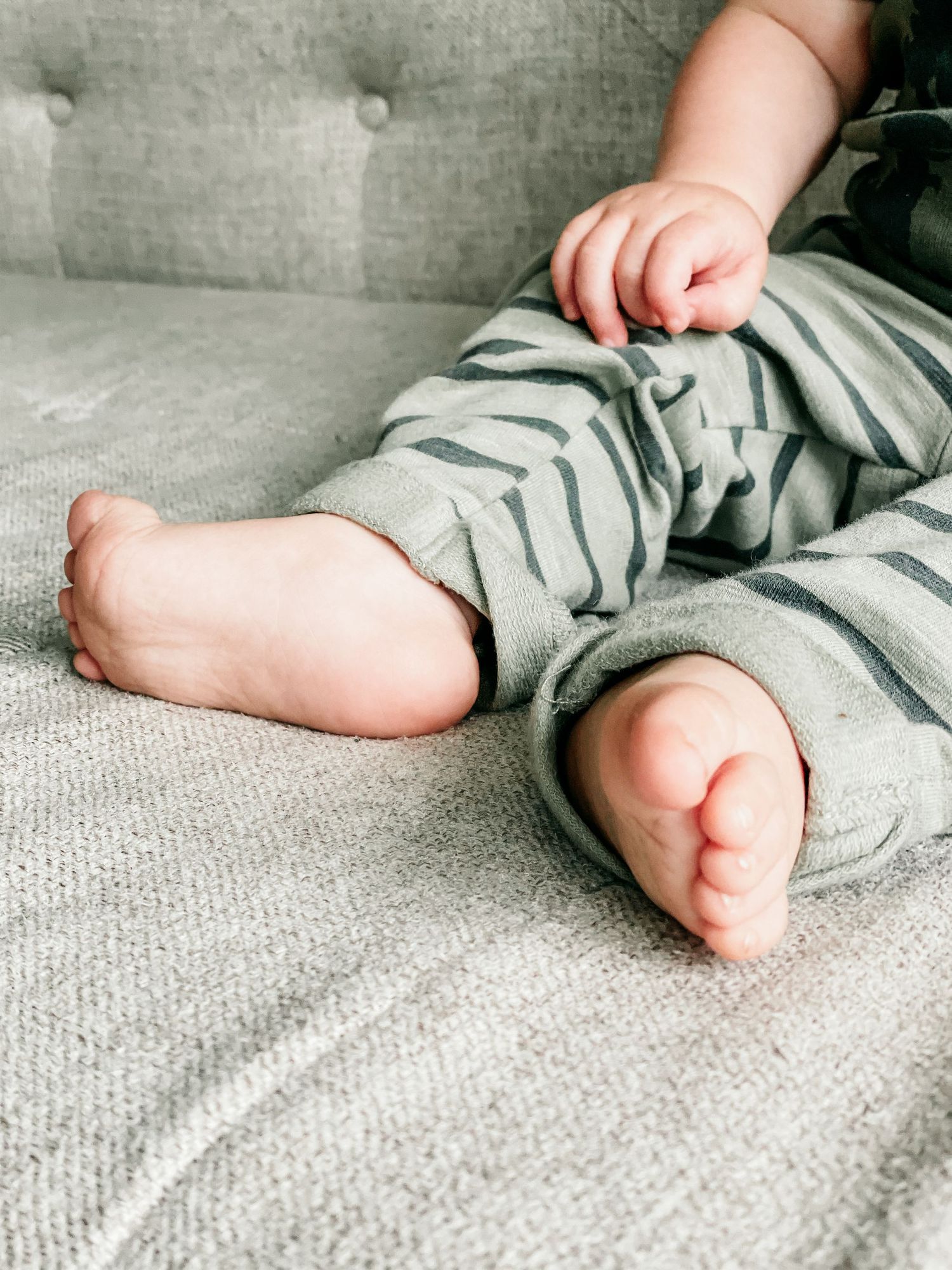 Close-up of baby feet and hands while sitting on a gray couch, wearing soft green and gray striped pajamas