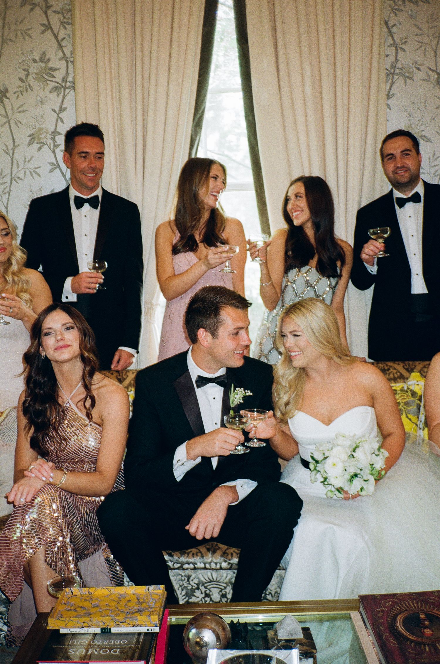 Wedding party guests in formal attire gather at elegant reception, raising champagne glasses in celebration.