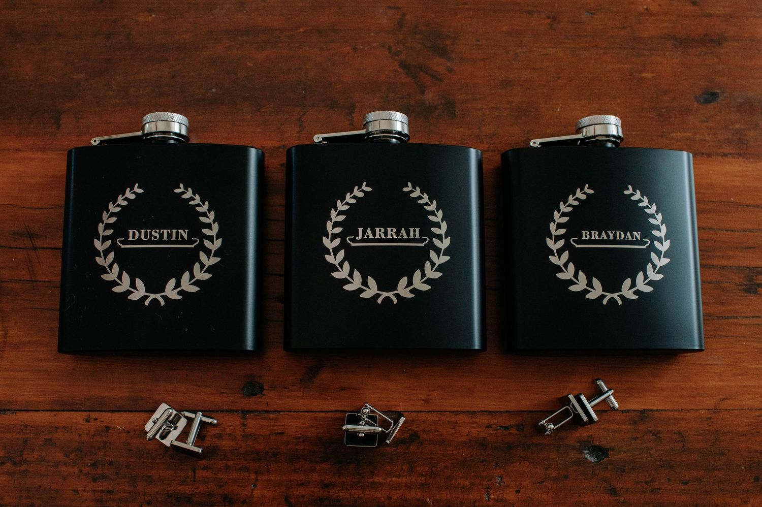 Three matching black flasks with laurel wreath designs and personalized names displayed beside cufflinks on wooden surface.