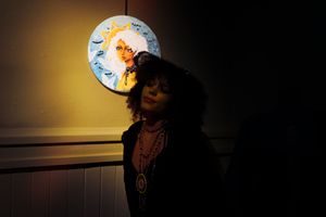 A person wearing Indigenous jewelry stands in dramatic lighting with a glowing circular artwork in the background.