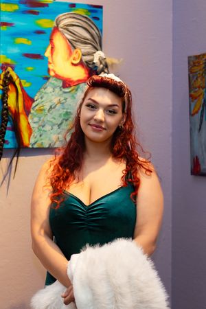 Woman in green velvet dress poses with white fur muff against colorful wall art display.