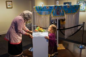 A masked worker stands behind a plexiglass barrier interacting with a young visitor at what appears to be a museum exhibit.