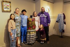 A group poses together at a clothing display in a retail or museum space with vintage dresses and outfits visible.