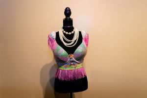 A mannequin displays a colorful pink and turquoise dance costume with silver necklaces against a beige background.