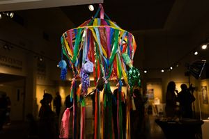 Colorful hanging art mobile with ribbons and decorative elements glowing in a dimly lit gallery space.