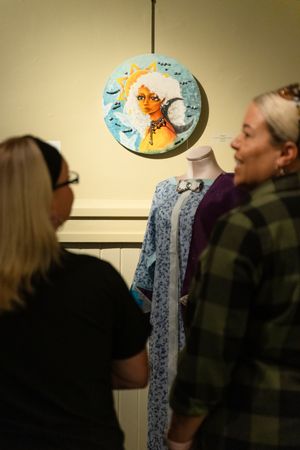 Viewers look at colorful circular artwork depicting sun and moon motifs hanging on beige wall.