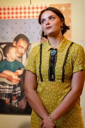 A person in a mustard yellow patterned dress and turquoise pendant stands thoughtfully against a wall with artwork.