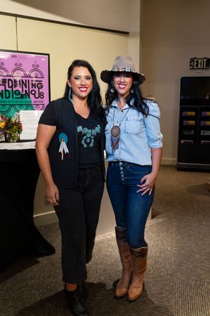 Two people pose together at cultural event, one wearing black shirt and one in denim with cowboy hat.