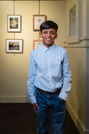 A youthful student wearing a blue gingham shirt stands in front of framed artwork.