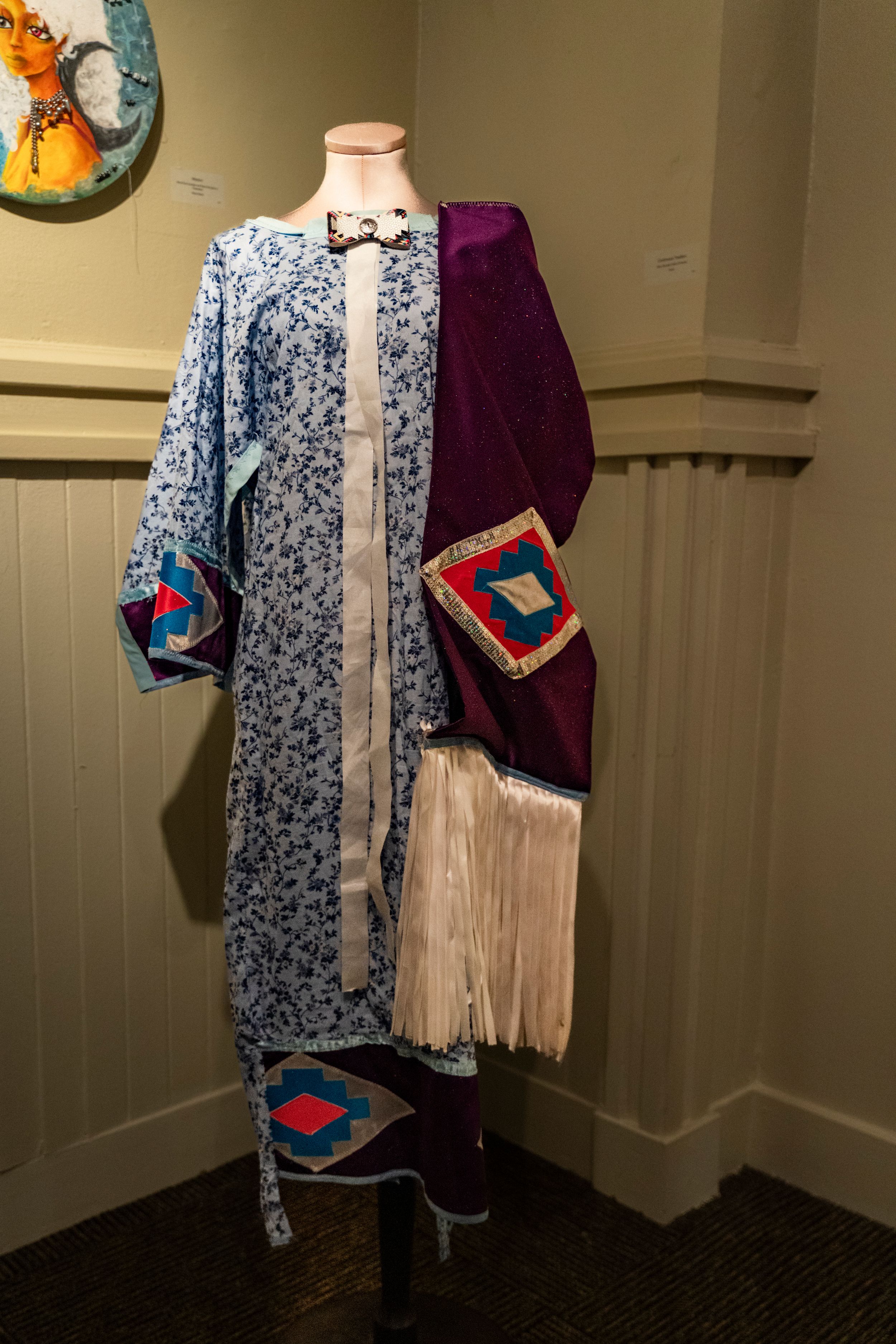 Traditional Native American dress with blue floral pattern, burgundy velvet panels, and geometric designs displayed on mannequin.