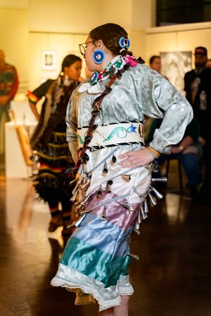 A performer wearing an ornate silver and teal regalia with fringe details dances at an indoor cultural event.