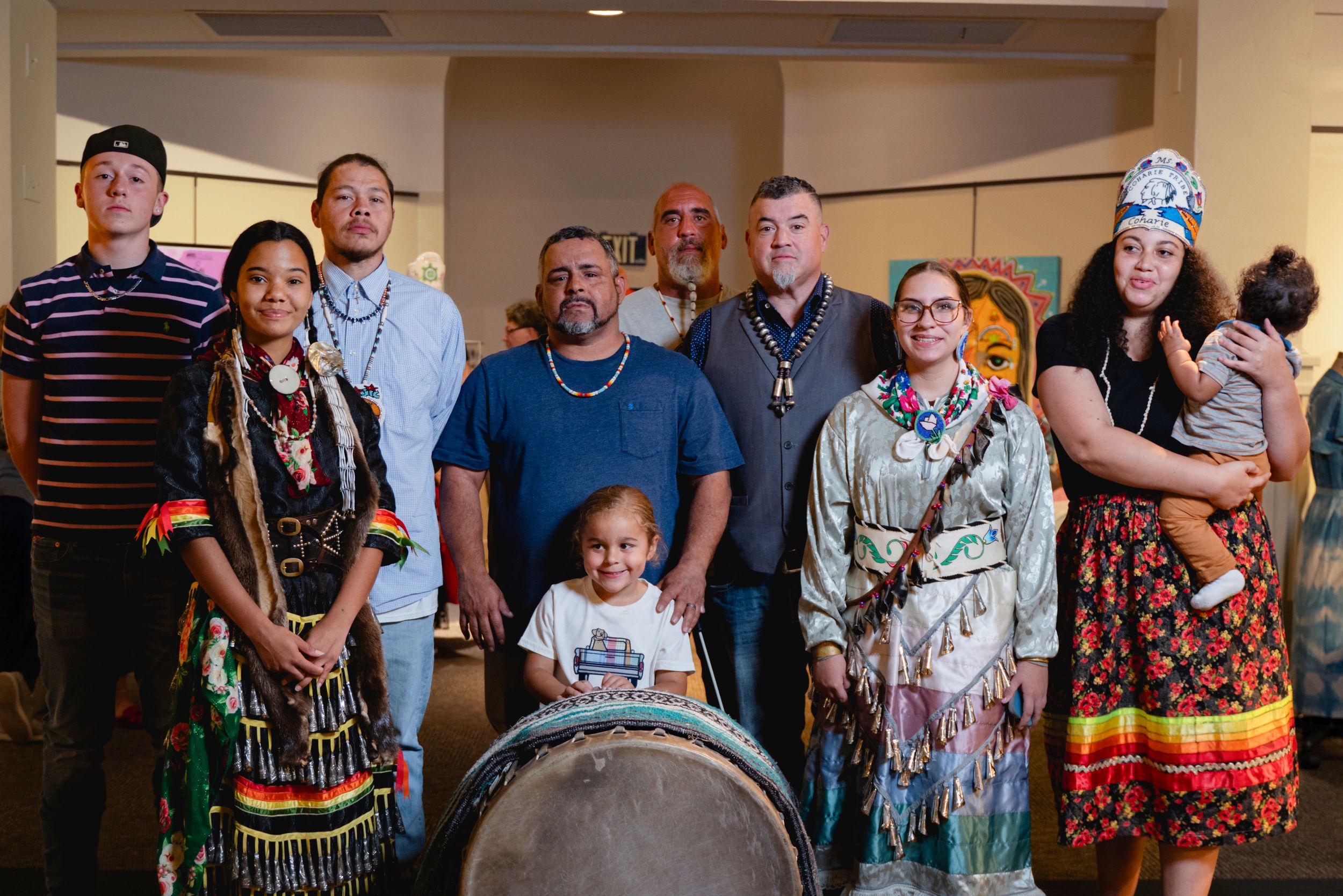 A diverse group in traditional regalia gather around a large drum in an indoor setting.