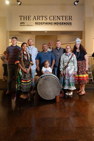 A group stands at The Arts Center under a 'Redefining Indigenous' sign with traditional dress and a large drum.