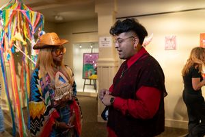 Two people in colorful outfits chat at an indoor event, with one wearing a vibrant multicolored ensemble.