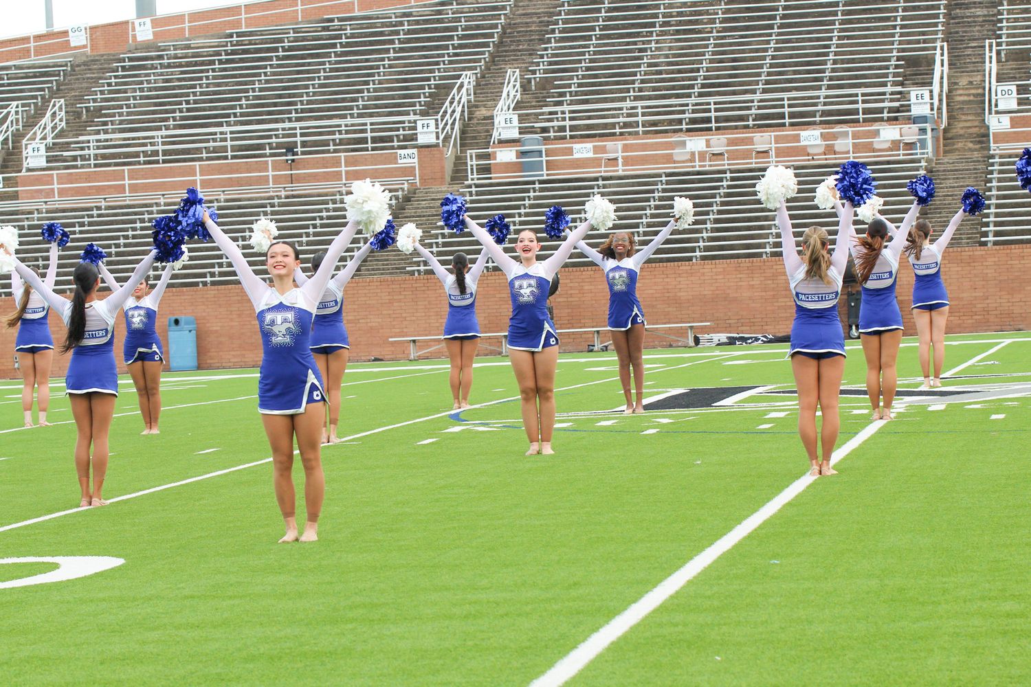 Cheerleaders in blue and white uniforms perform stunts and routines on a football field with stadium bleachers behind them.