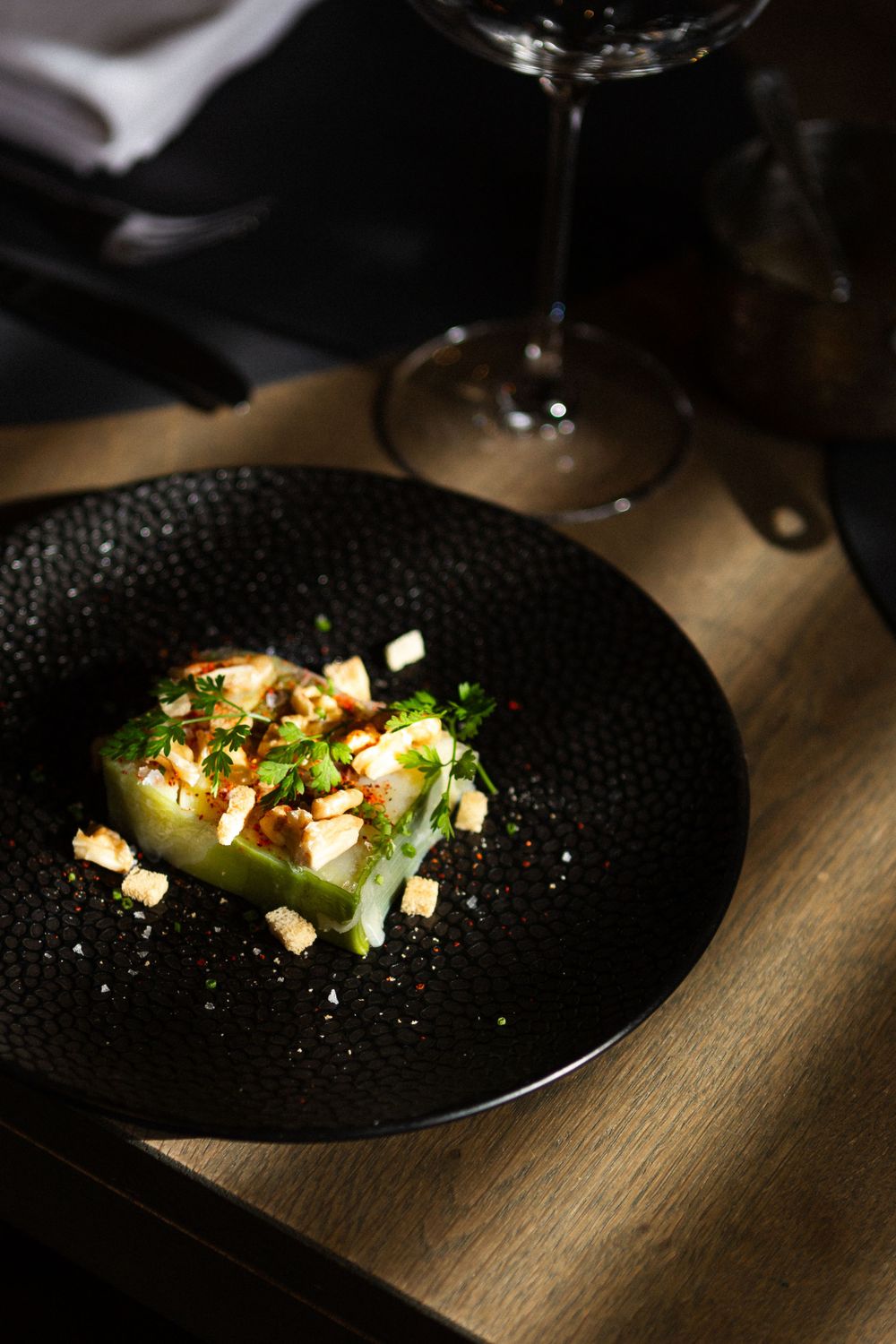 Elegant plating of a light green dish garnished with nuts and herbs on a black plate with wine glass in background.