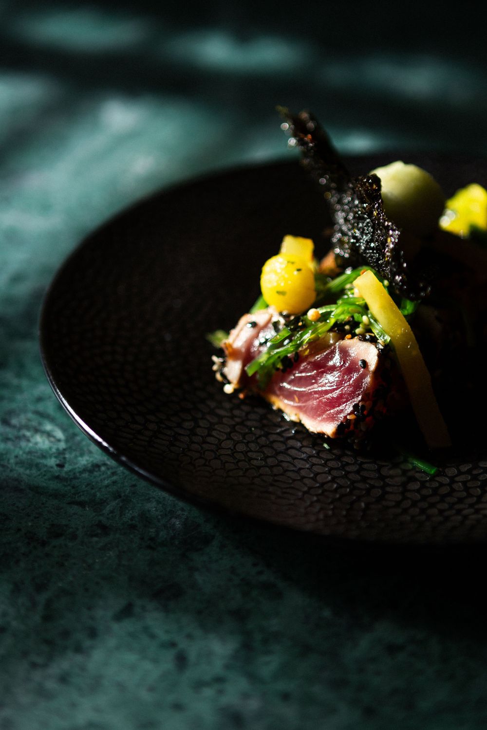 Seared tuna with yellow mango cubes and microgreens served on a textured black plate against teal background.