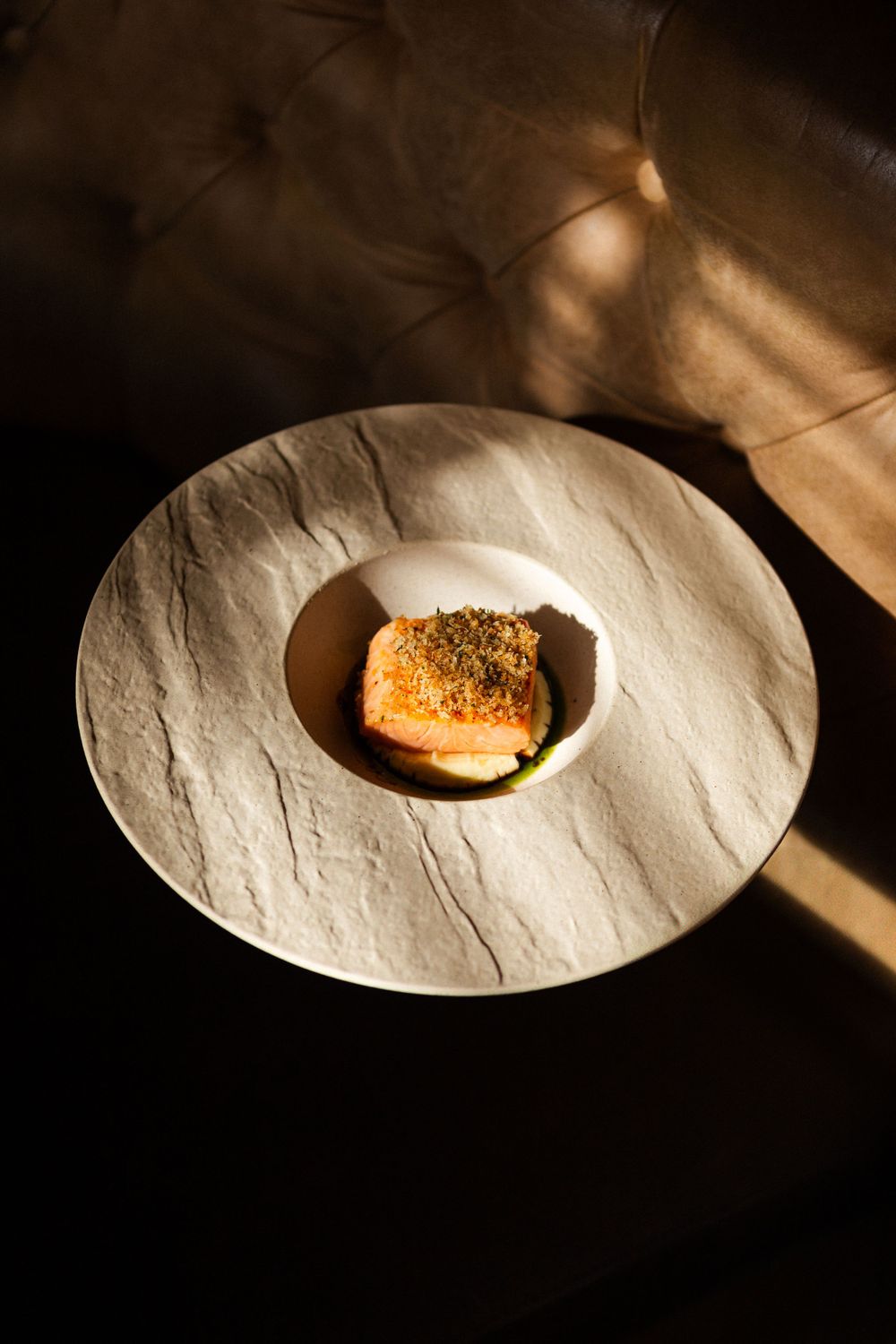 A piece of cooked salmon with crispy seasoned topping served on a textured white plate in dramatic lighting.