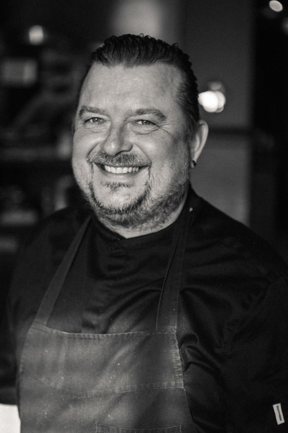 Black and white portrait of a smiling chef wearing a dark apron and chef coat in a professional kitchen setting.