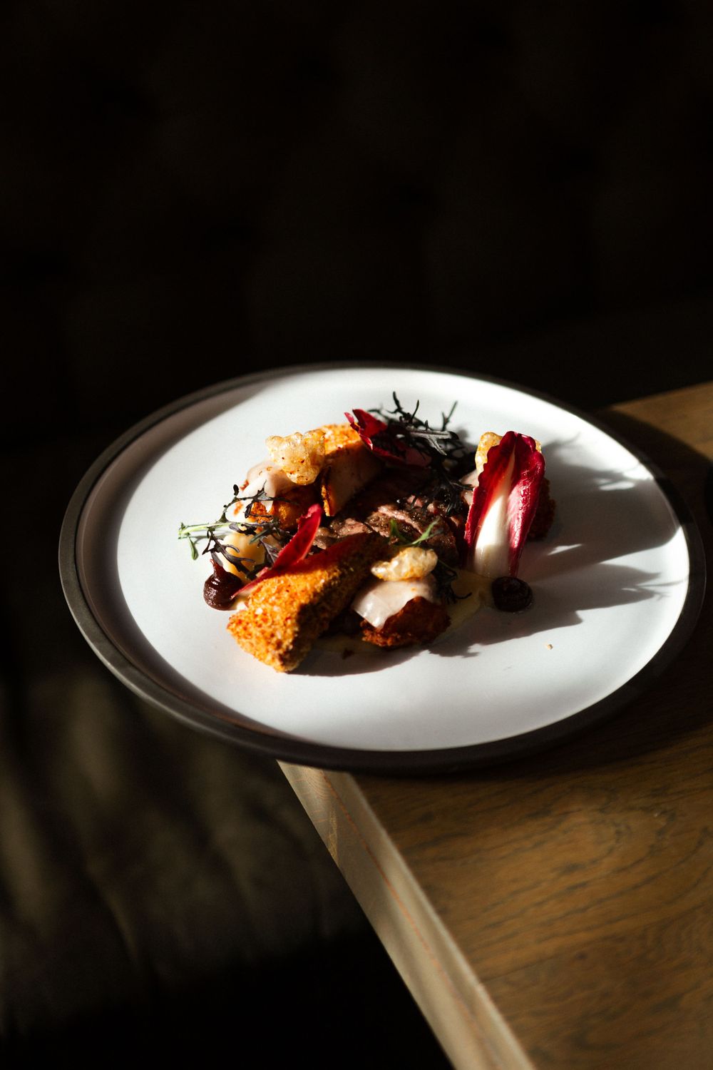 Elegant plated dish with dark meat, vibrant citrus, and herbs cast in dramatic lighting on white ceramic plate.