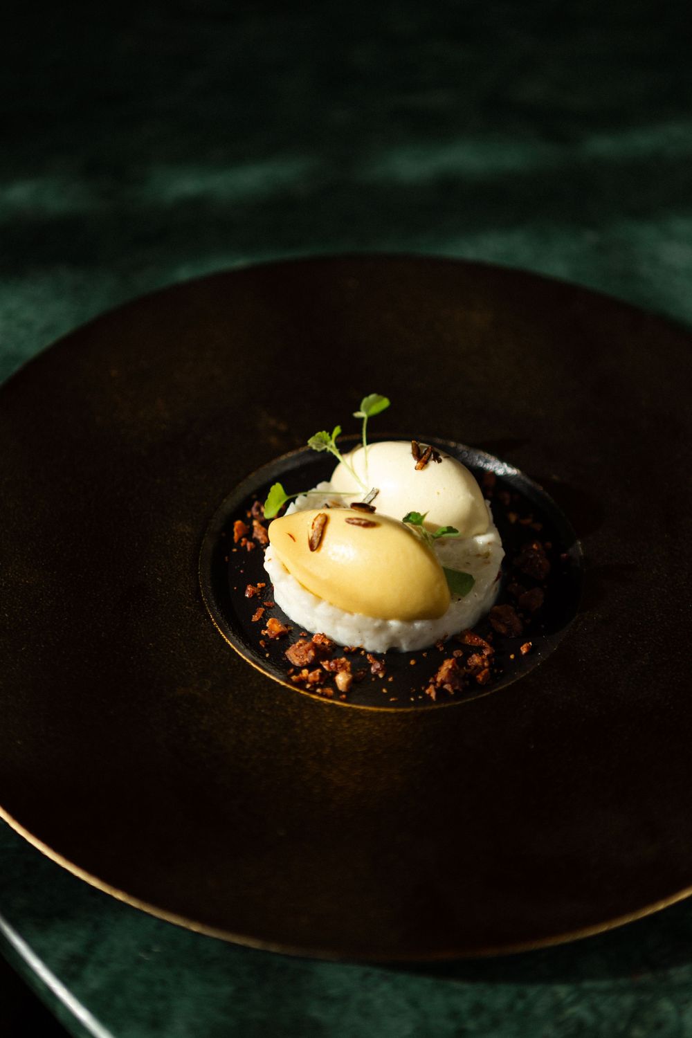 A plated dessert with vanilla ice cream and poached pear on a dark chocolate crumb base presented on a black plate.