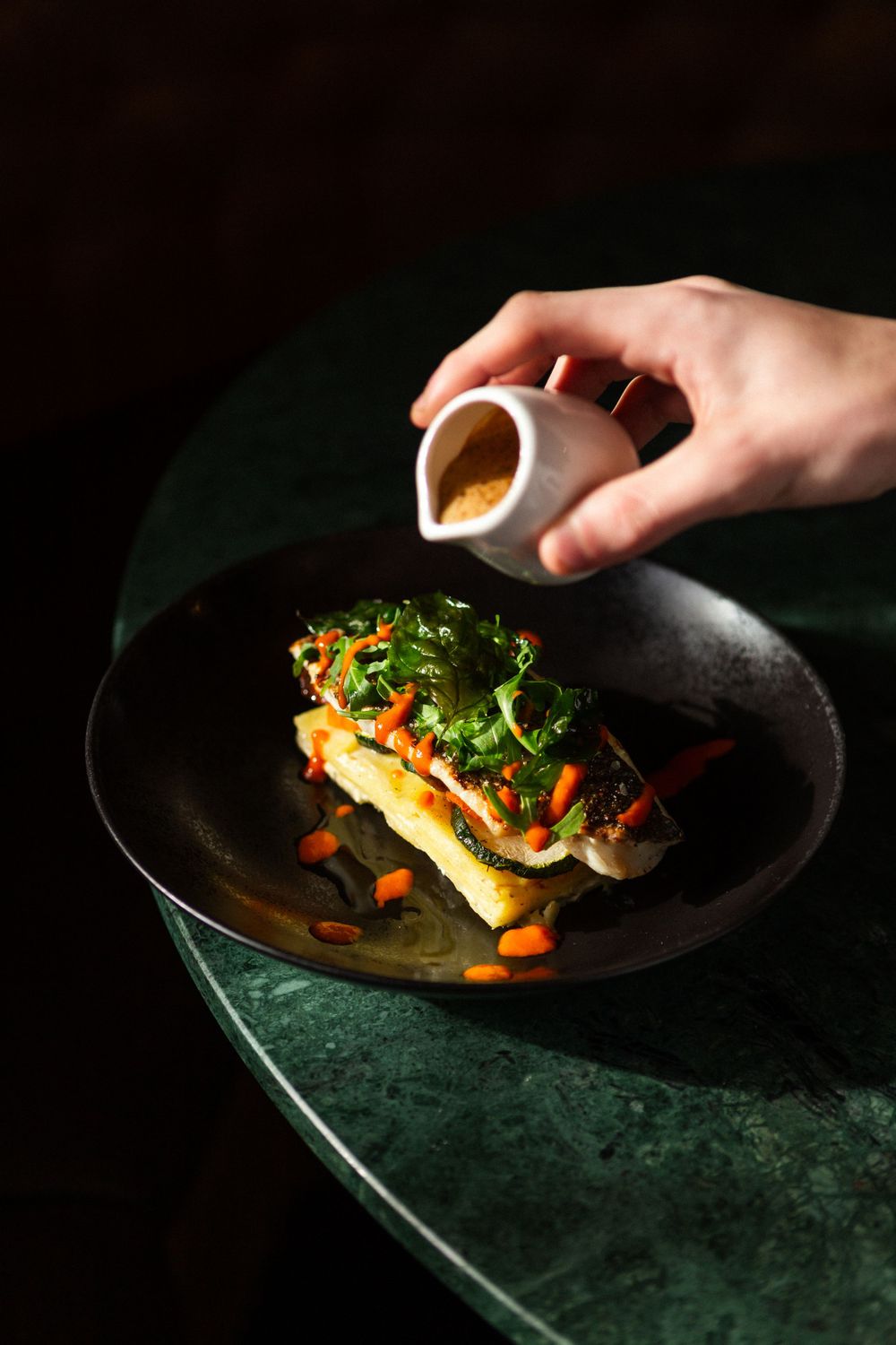 Elegant plated fish dish with microgreens and vegetables being finished with a drizzle of sauce in dramatic lighting.