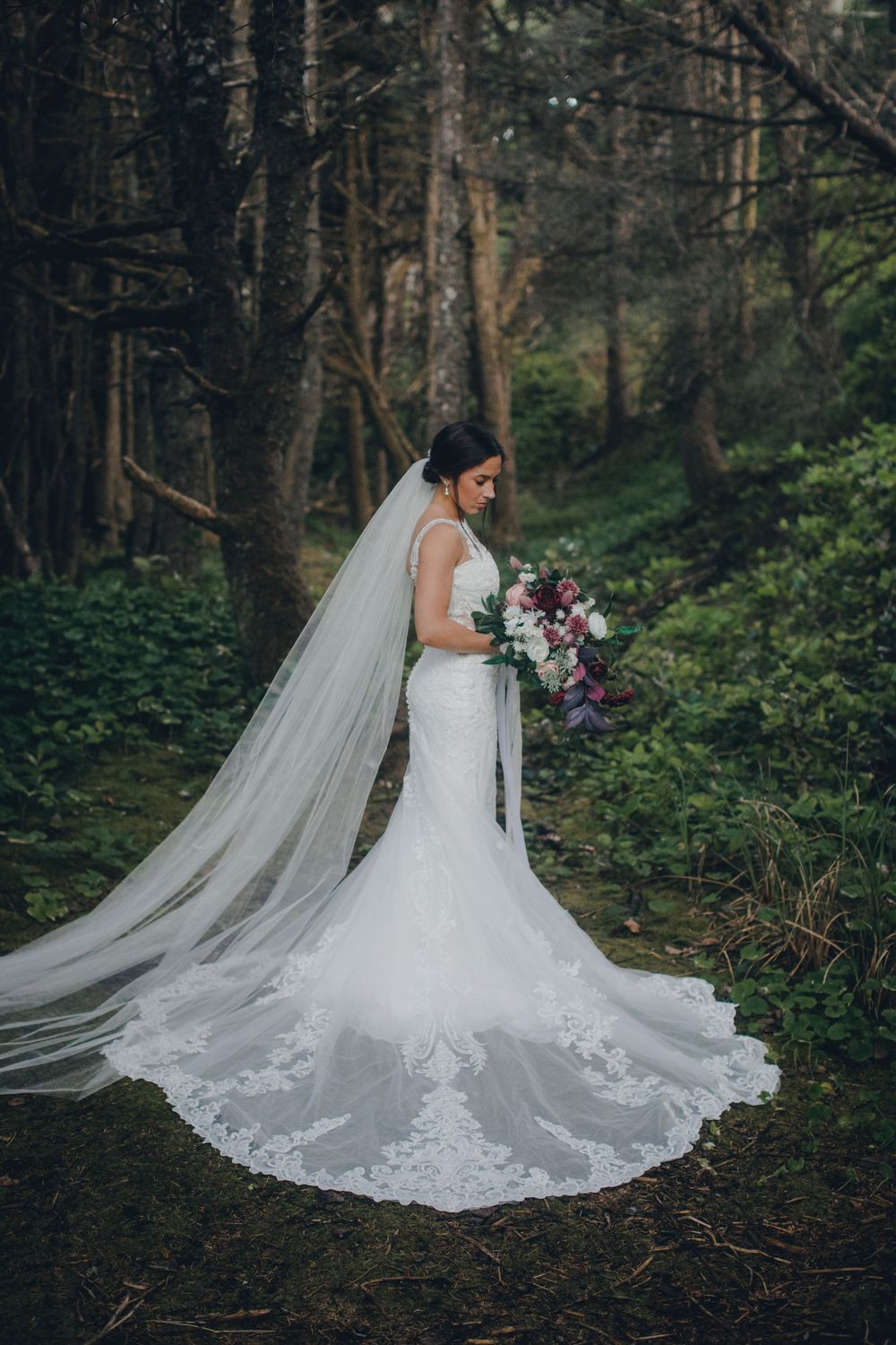 A bride in a lace mermaid wedding dress with long flowing veil poses in a moody forest setting holding a purple bouquet.