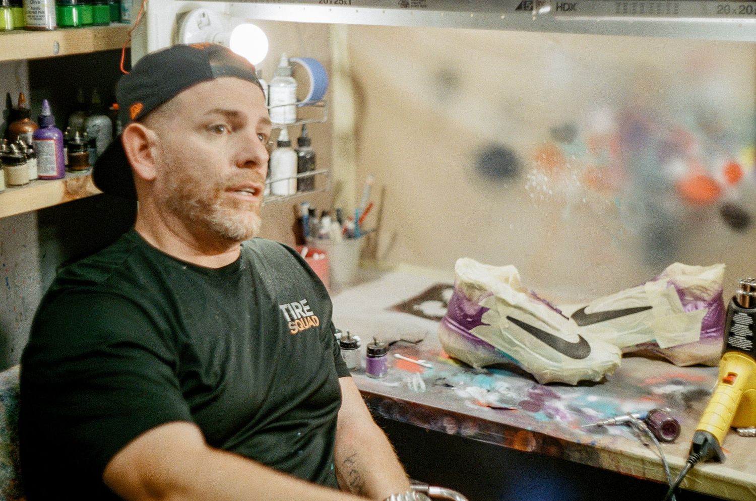 Craftsman wearing a black t-shirt sitting in a workshop with tools and supplies on cluttered workbench.