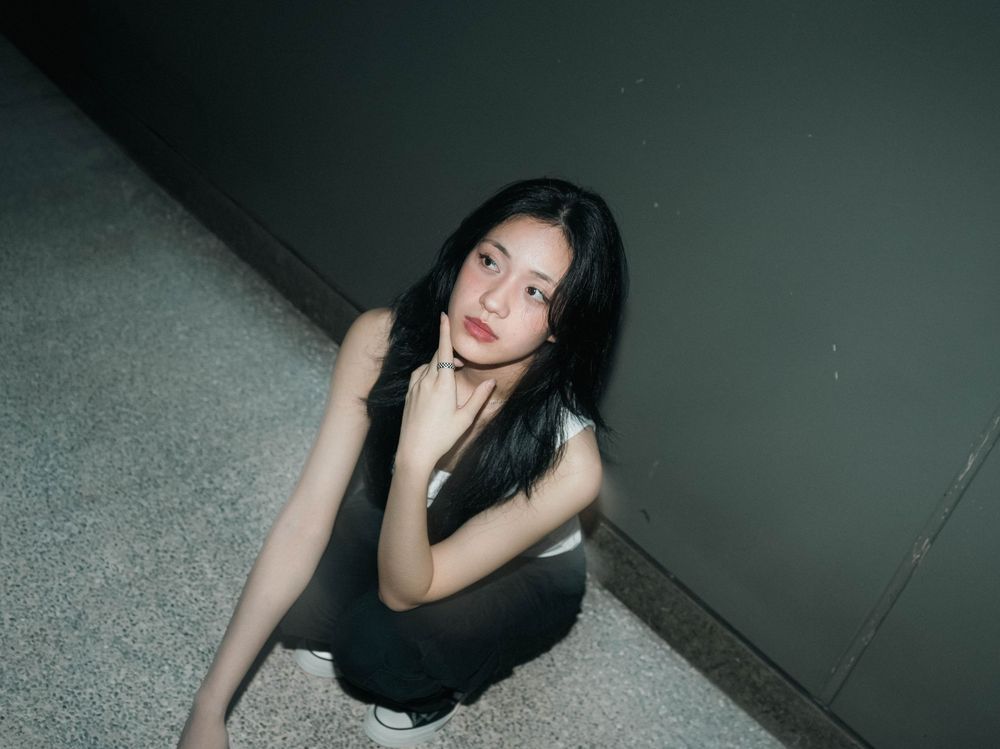 A person in a black outfit sits crouched against a dark wall in moody lighting.