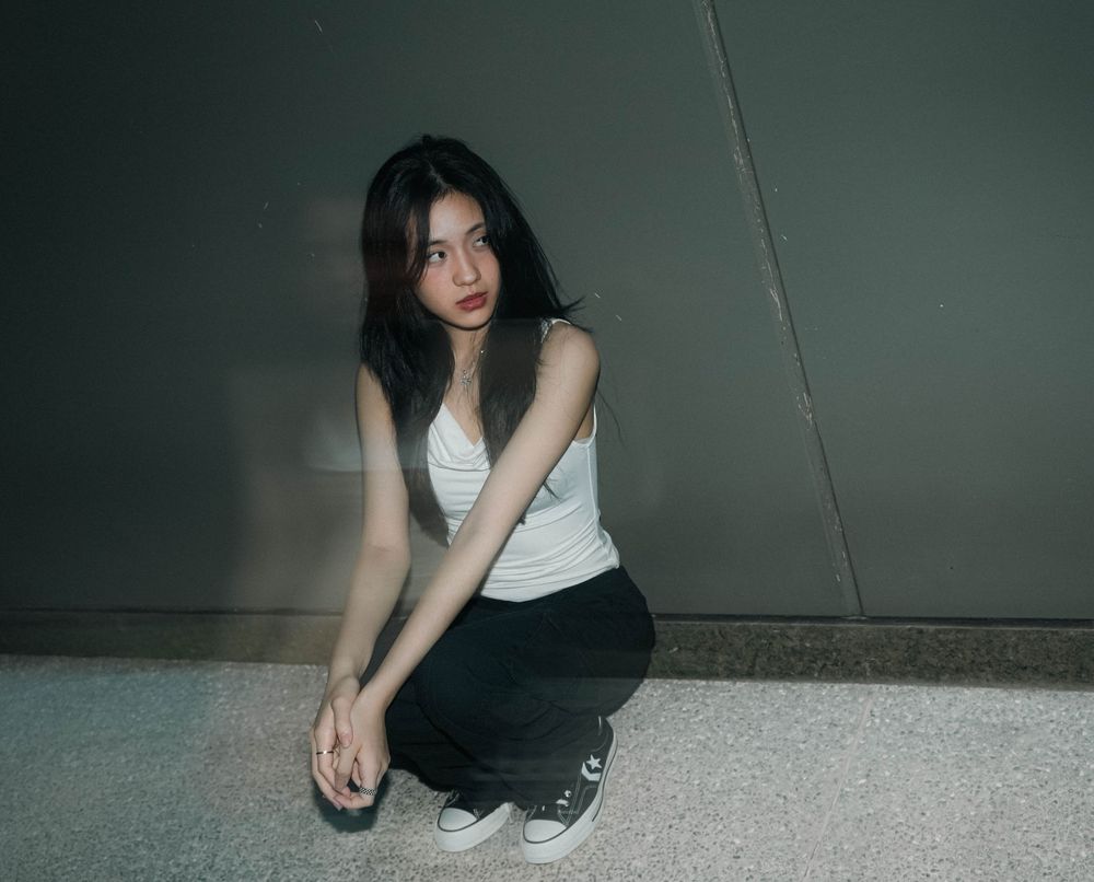 Someone in a casual white tank top and black pants crouches on the ground in a dimly lit hallway.