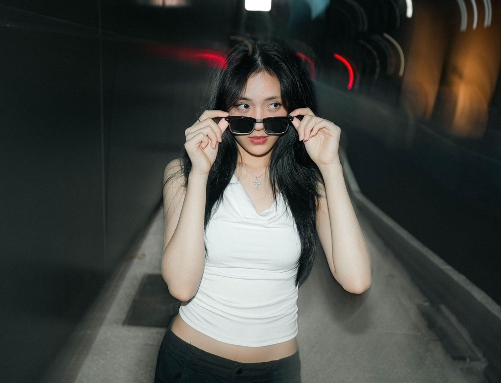 Woman in a white tank top lifting dark sunglasses with both hands while standing in a dimly lit tunnel.