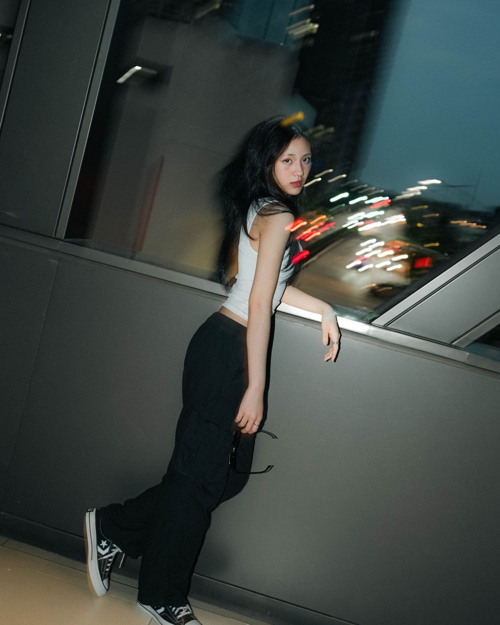 Figure leaning against wall in dark atmospheric setting with blurry lights in background wearing casual attire.