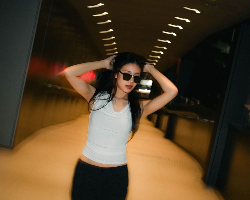 Person wearing sunglasses and crop top poses energetically in a tunnel with fluorescent lighting overhead.