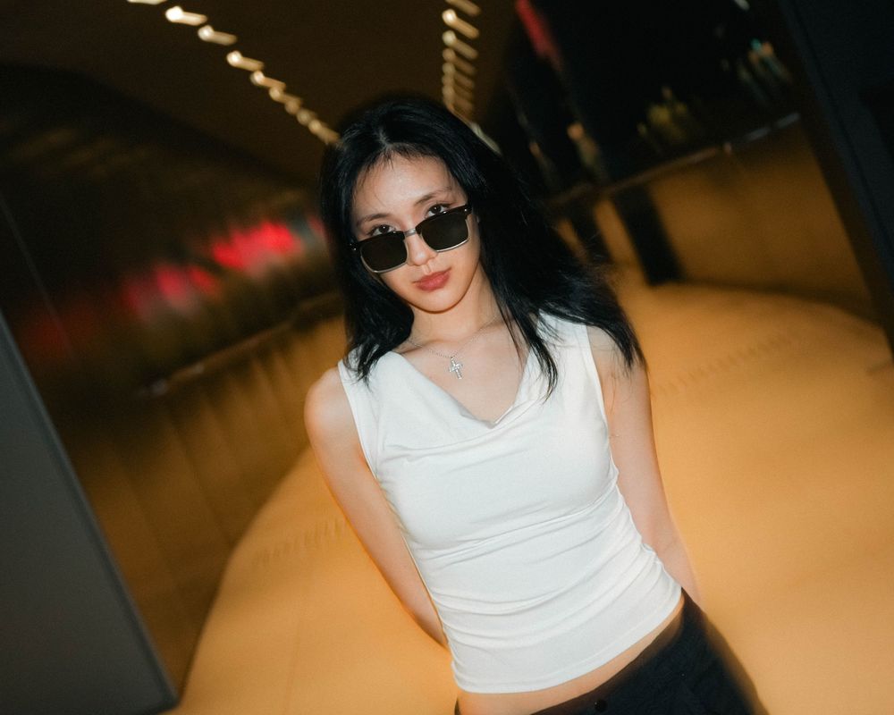 Person in white crop top and sunglasses poses in a dimly lit setting with dramatic lighting effects.