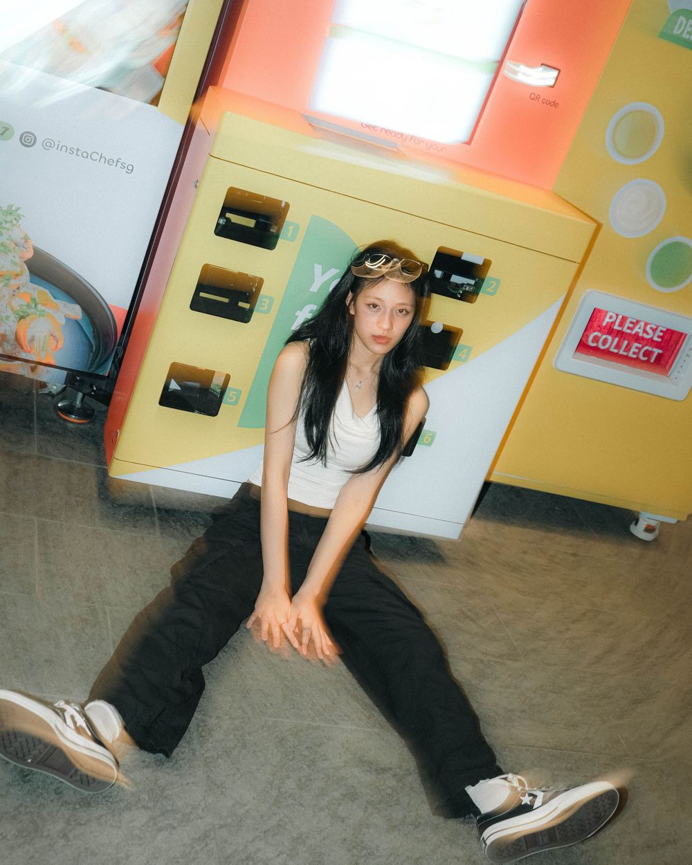 Person in casual attire with sunglasses on head sits on floor against colorful wall with lockers in urban setting.