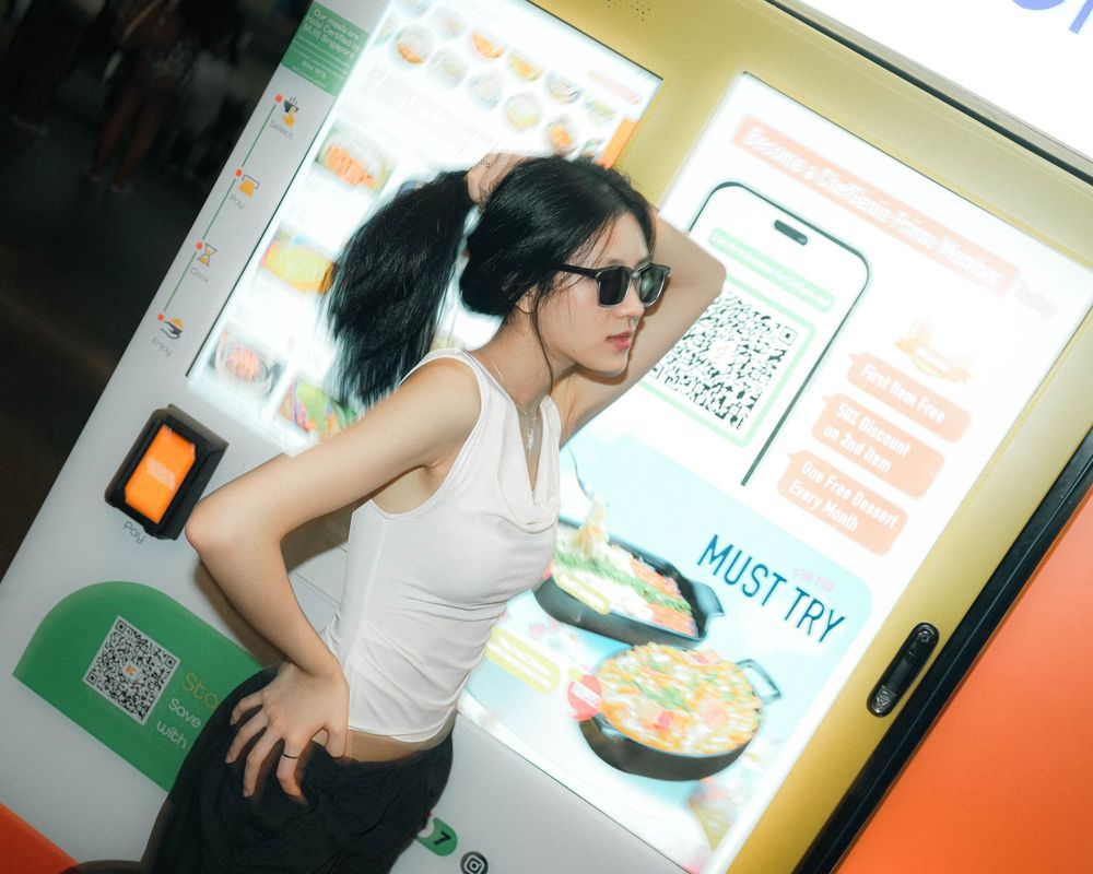 A customer in sunglasses and white tank top looks at a digital menu screen at a restaurant kiosk.