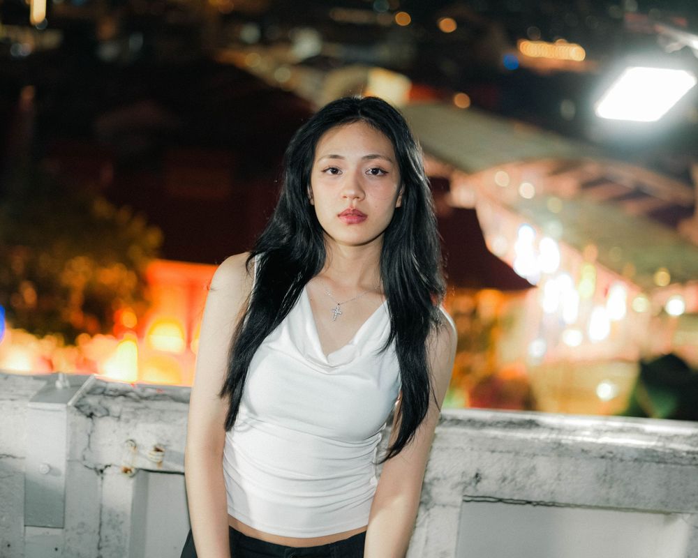 A woman with long dark hair poses at night on a rooftop overlook with city lights in the background.