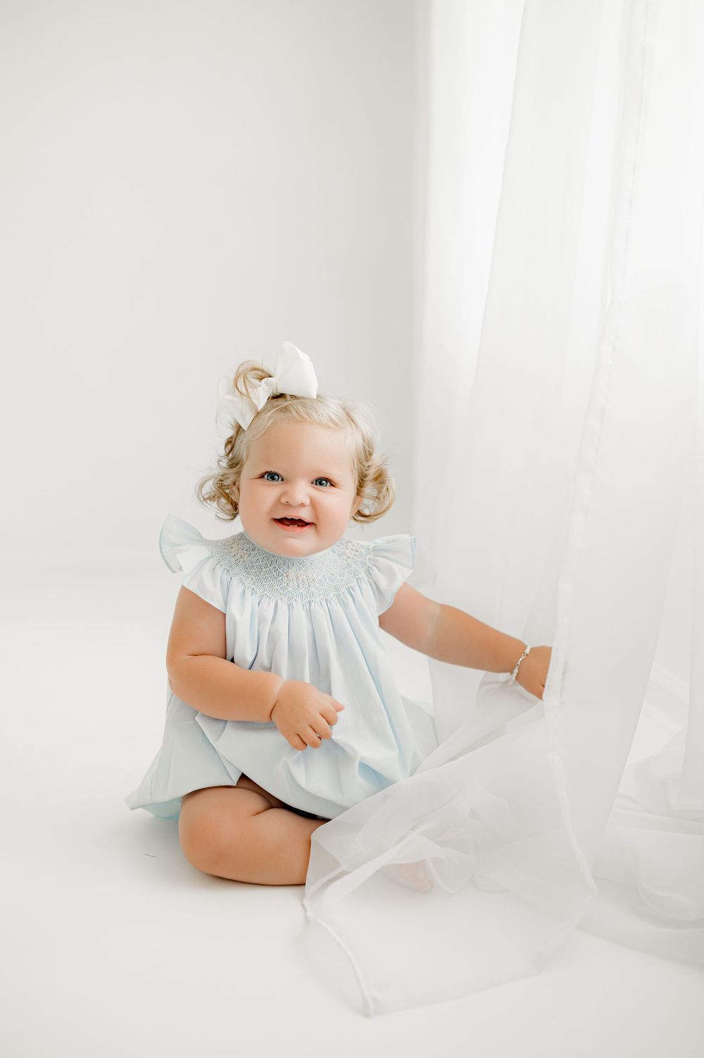 Adorable baby in light blue dress and bow smiles while playing with sheer curtains in white studio.