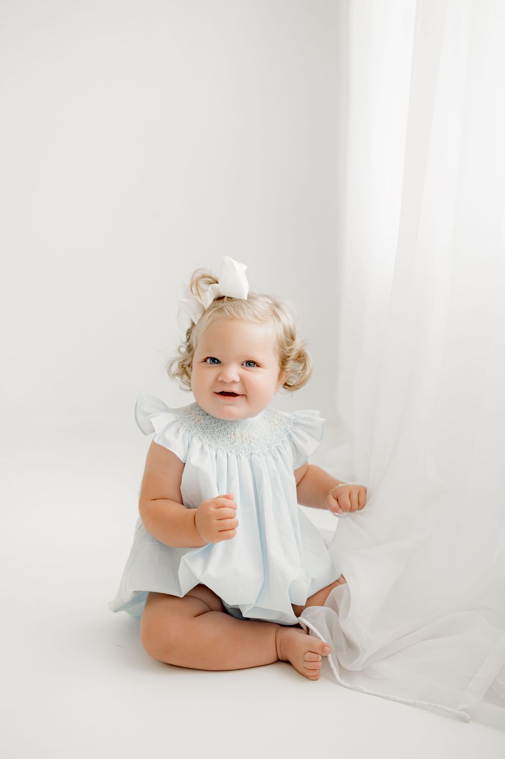 A joyful toddler in a white dress and bow sits against a minimalist white background.