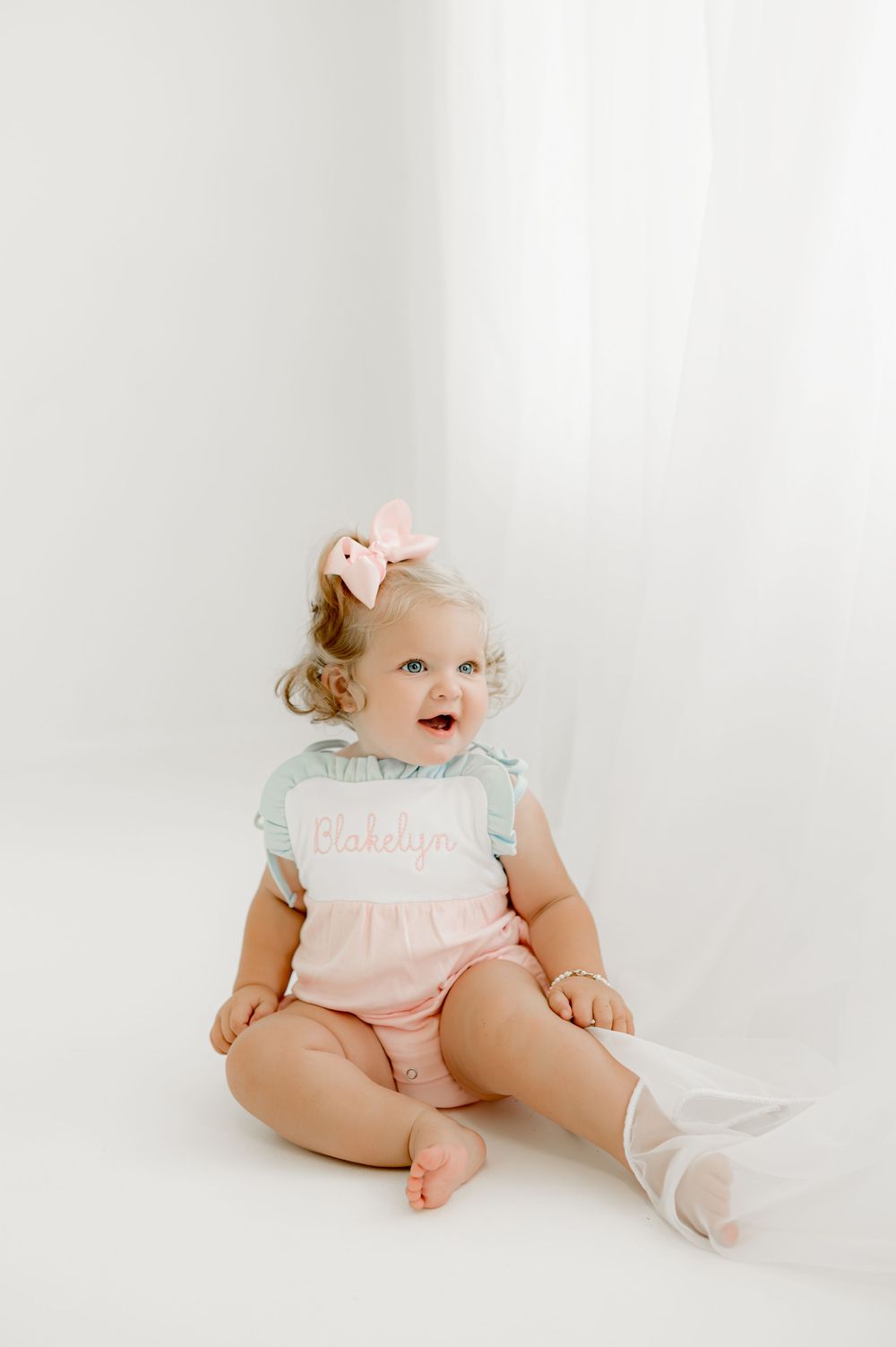 Baby in white and peach outfit laughs while sitting on white studio floor.