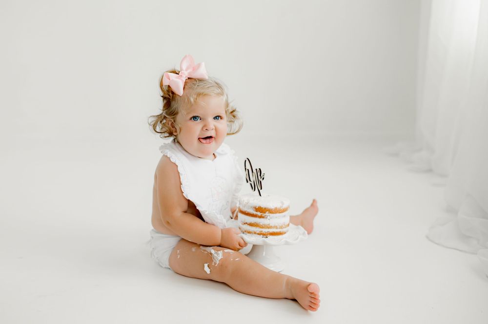 Infant sitting on white background wearing bodysuit with pink bow in hair during first birthday cake smash session.