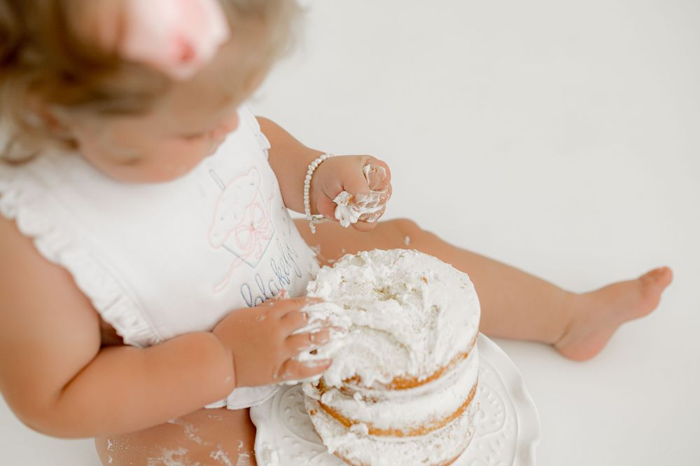 Baby in white dress holding and playing with frosted cake during cake smash photoshoot.