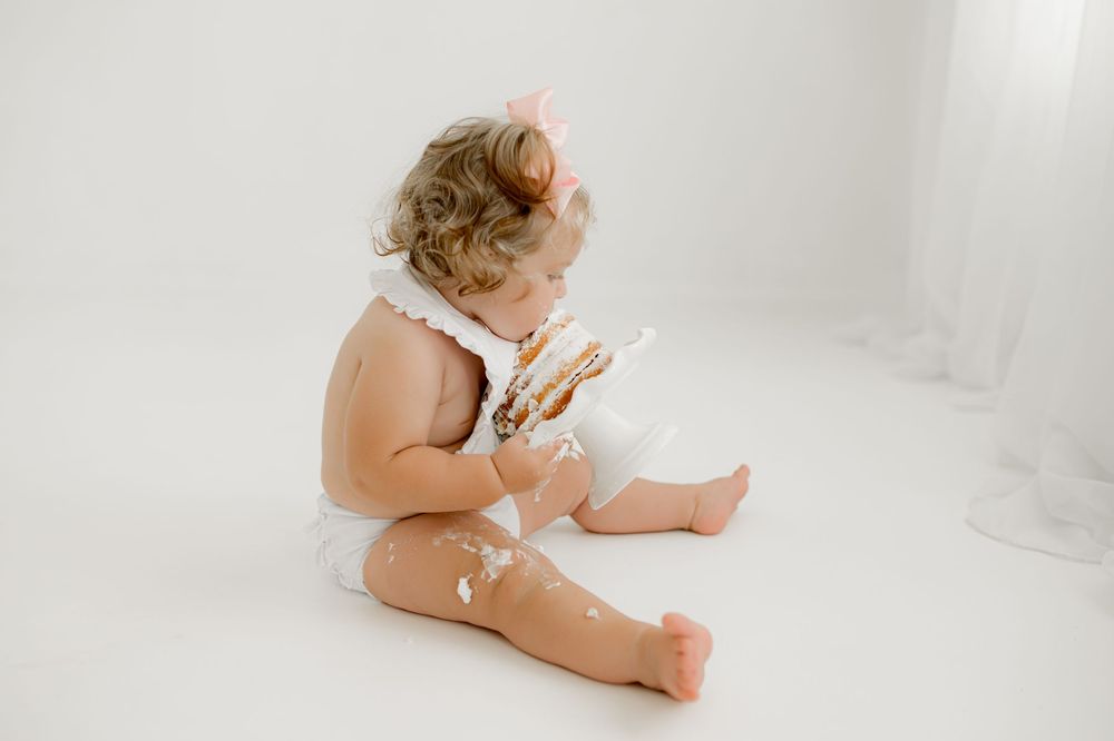Baby in white lace bodysuit sits barefoot on clean white backdrop during studio photo session.